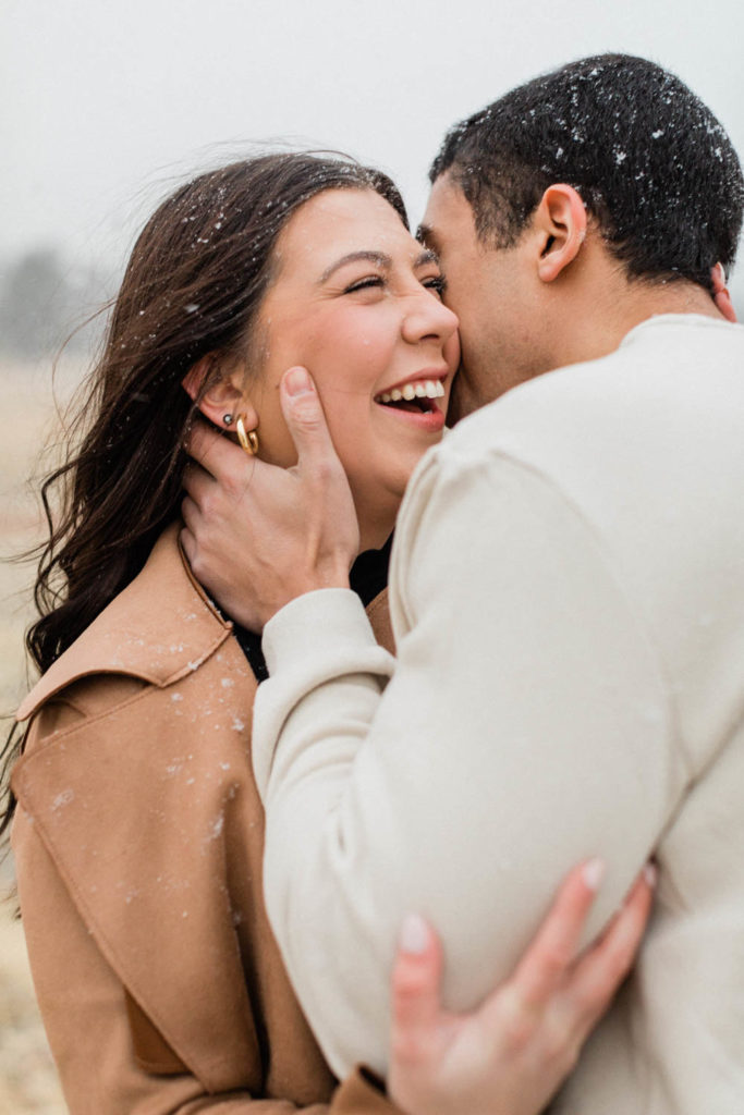 guy whispering into his girlfriends ear and making her laugh while holding her face during their snowy Boulder engagement session holding hands with the Colorado Rocky Mountains behind them with Colorado elopement photographer Diana Coulter
