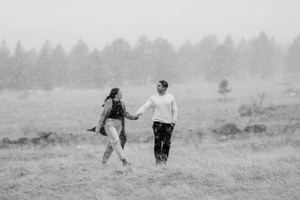 stylish couple walking through the field during their snowy engagement session in the Boulder Flatirons holding hands with the Colorado Rocky Mountains behind them with Colorado elopement photographer Diana Coulter