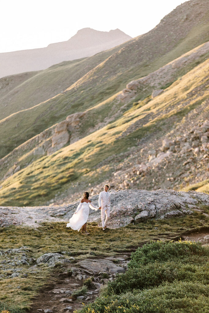 biracial couple walking in the mountains in their wedding day attire at sunrise looking off into the distance