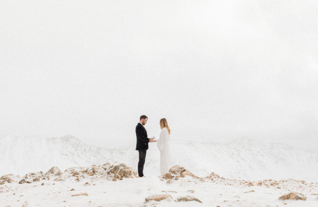 bride holding grooms hand as he reads his vows to her while its snowing on the top of a mountain in colorado in the winter