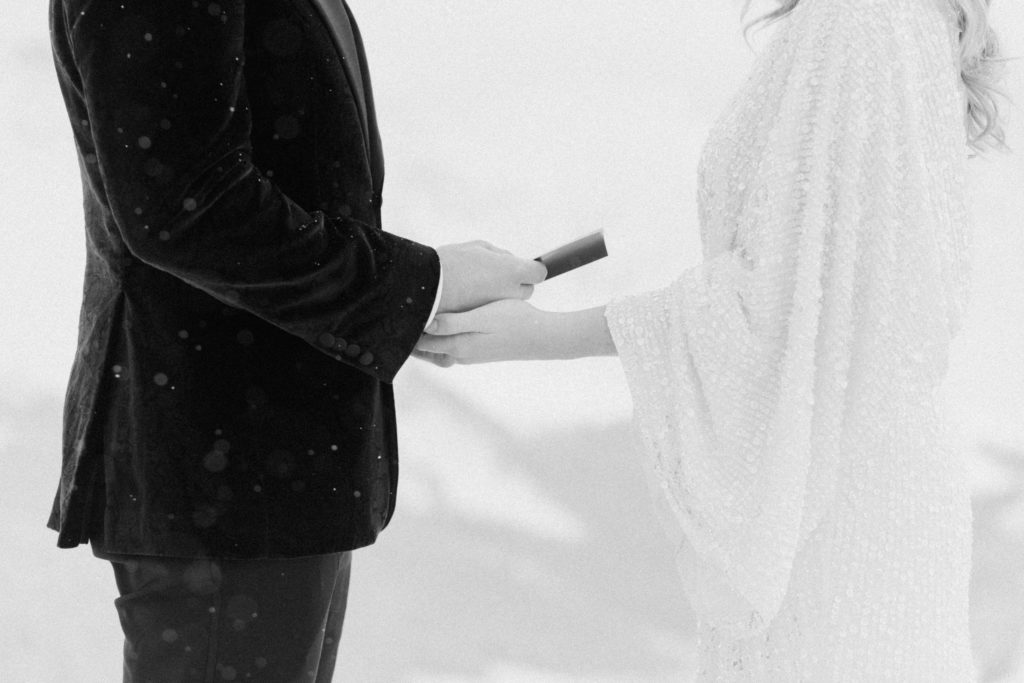 black and white close up of bride holding grooms hand while he reads his wedding vows to her during magical snowfall at the top of a mountain in colorado during elopement 