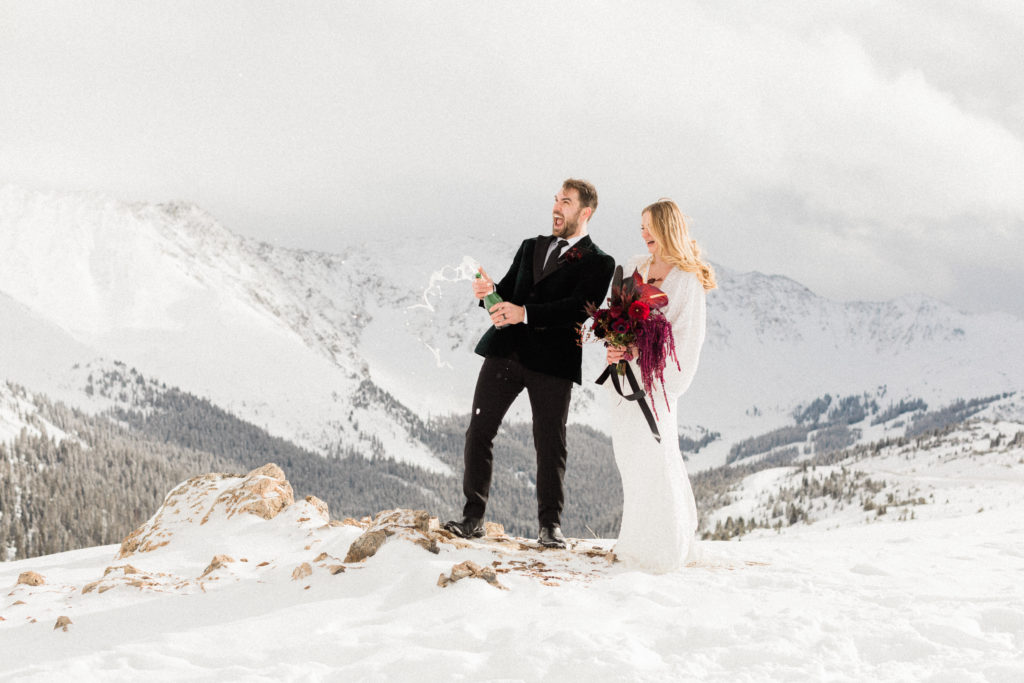 bride and groom poppin champagne at the top of Loveland pass after reading vows and eloping in Colorado as bride carries her bouquet from denver florist project floral