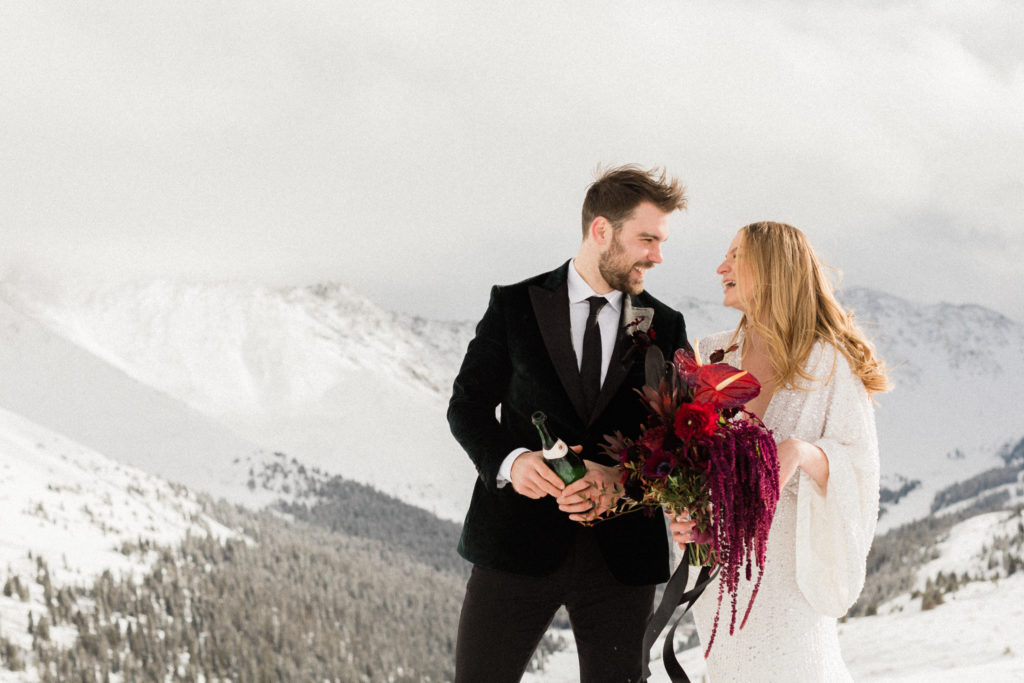 bride and groom poppin champagne at the top of Loveland pass after reading vows and eloping in Colorado as bride carries her bouquet from denver florist project floral