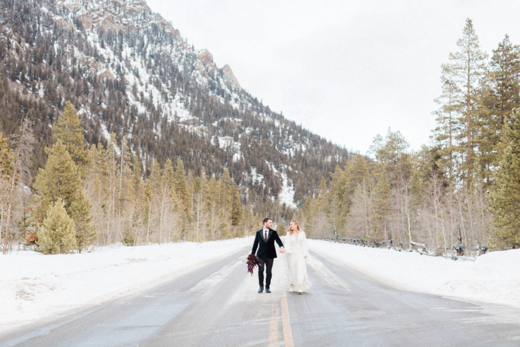 groom holding bride's moody red floral bouquet from the perfect petal as they walk in the street in the mountains in breckenridge colorado during their snowy winter elopement by Diana Coulter