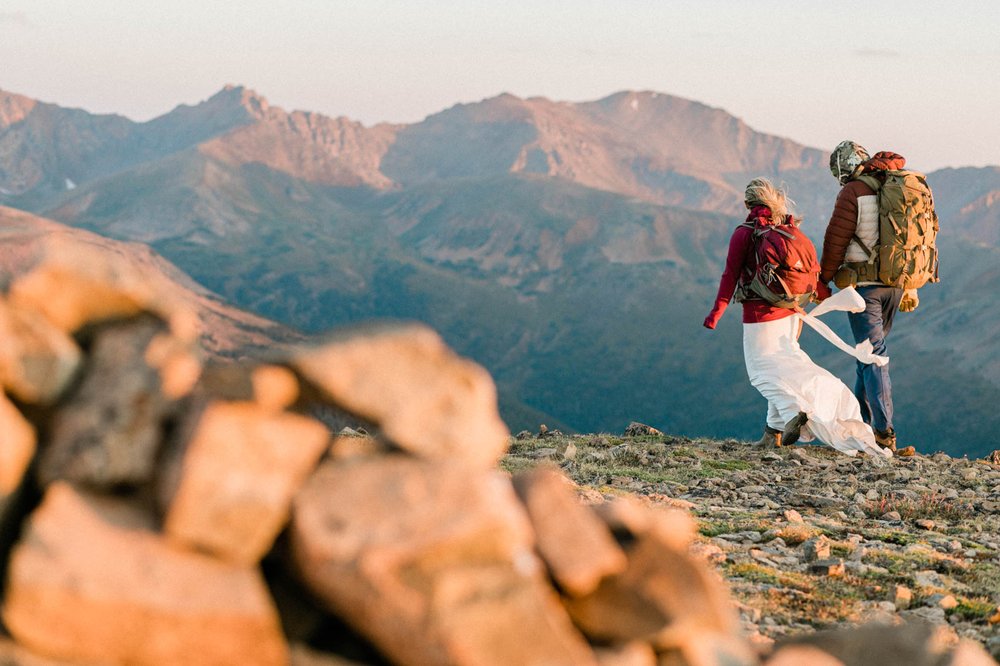 bride and groom walking on loveland pass during cold september morning bride wearing wedding dress and red coat, groom wearing hat tux coat alpenglow during summer sunrise adventure elopement at loveland pass near breckenridge, colorado