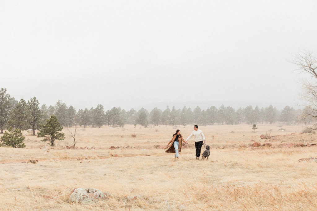 stylish couple walking through the field during their snowy engagement session in the Boulder Flatirons holding hands with the Colorado Rocky Mountains behind them with Colorado elopement photographer Diana Coulter