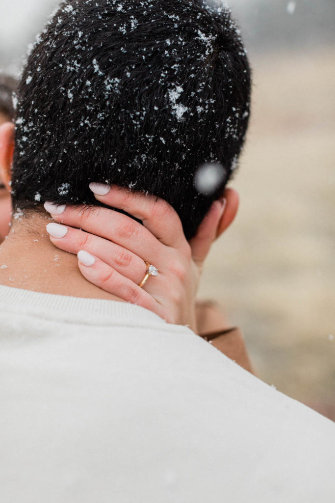 close up photo of engagement ring with girl's hand wrapped around her fiance's neck while pulling him into kiss him during snowy engagement session in boulder colorado by diana coulter