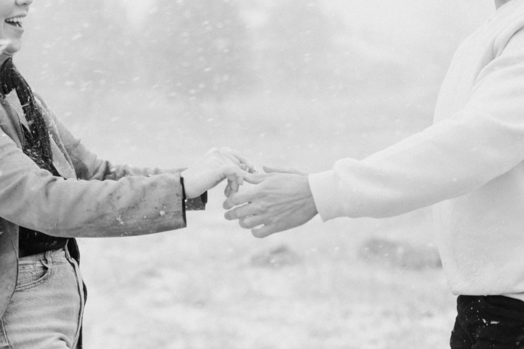 Black and white photo of couple holding hands and dancing in the mountains during their playful engagement session in the snow in Boulder, Colorado with wedding photographer Diana Coulter