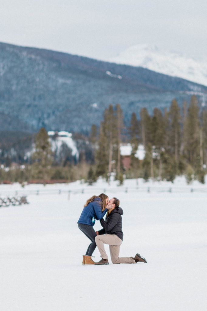 boyfriend down on one knee proposing to girlfriends during secret surprise proposal at devil's thumb ranch in the winter on new years eve with snow on the ground. bride is leaning down to say yes and kiss the groom. they are wearing Patagonia jackets because it's cold out and Sorel boots to keep their feet warm.
