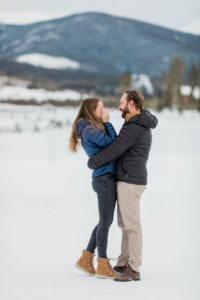 groom is holding bride's waist while bride cover's her eyes because she is crying out of sheer happiness, joy and shock during secret surprise proposal at devil's thumb ranch in the winter on new years eve with snow on the ground. they are wearing Patagonia jackets because it's cold out and Sorel boots to keep their feet warm.