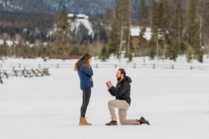 boyfriend down on one knee proposing to girlfriends during secret surprise proposal at devil's thumb ranch in the winter on new years eve with snow on the ground. bride is covering her eyes because she's crying. they are wearing Patagonia jackets because it's cold out and Sorel boots to keep their feet warm.