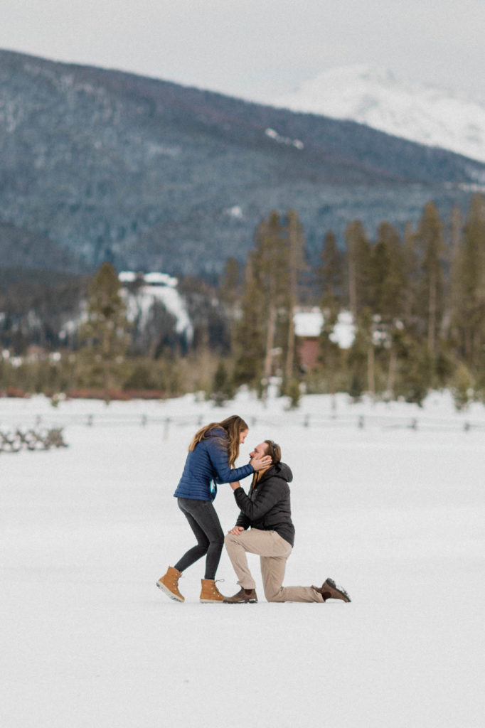 boyfriend down on one knee proposing to girlfriends during secret surprise proposal at devil's thumb ranch in the winter on new years eve with snow on the ground. bride is leaning down to say yes and kiss the groom. they are wearing Patagonia jackets because it's cold out and Sorel boots to keep their feet warm.
