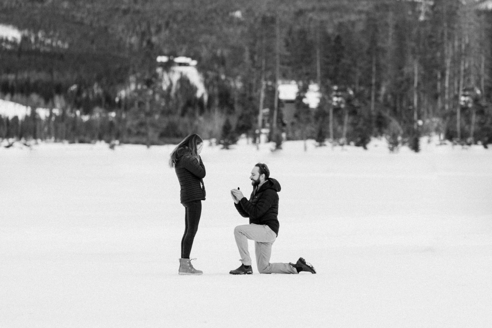 black and white photo of a man proposing to woman at devils thumb ranch on new years eve in the winter. He is down on one knee opening a ring box with a huge diamond ring on the inside asking her to marry him.
