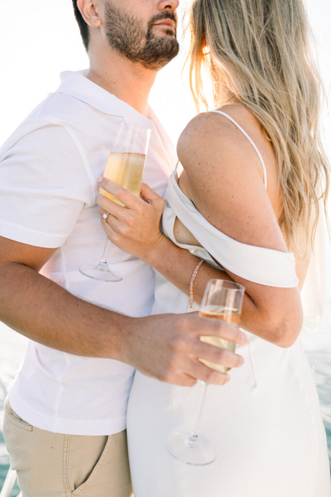 Couple standing together facing each other on a sailboat in Chicago holding glasses of Veuve Cliquot champagne during their sunrise engagement session on Lake Michigan