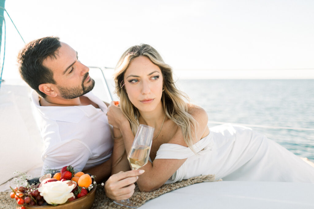 Couple laying down on a sailboat in Chicago holding glasses of Veuve Cliquot champagne while eating strawberries during their sunrise engagement session on Lake Michigan